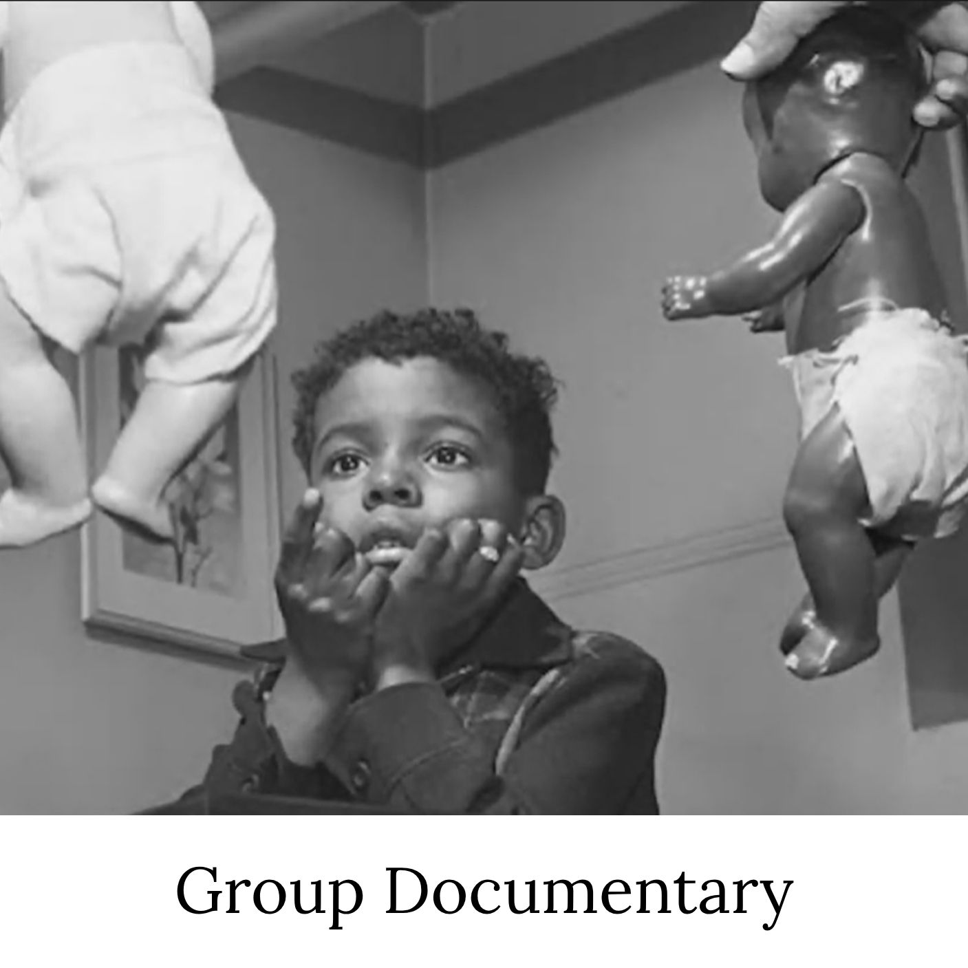 B&W photo showing two dolls with dark and light skin colors being presented to a Black child, whose attention is drawn to the doll with the paler skin. The image is labeled Group Documentary.