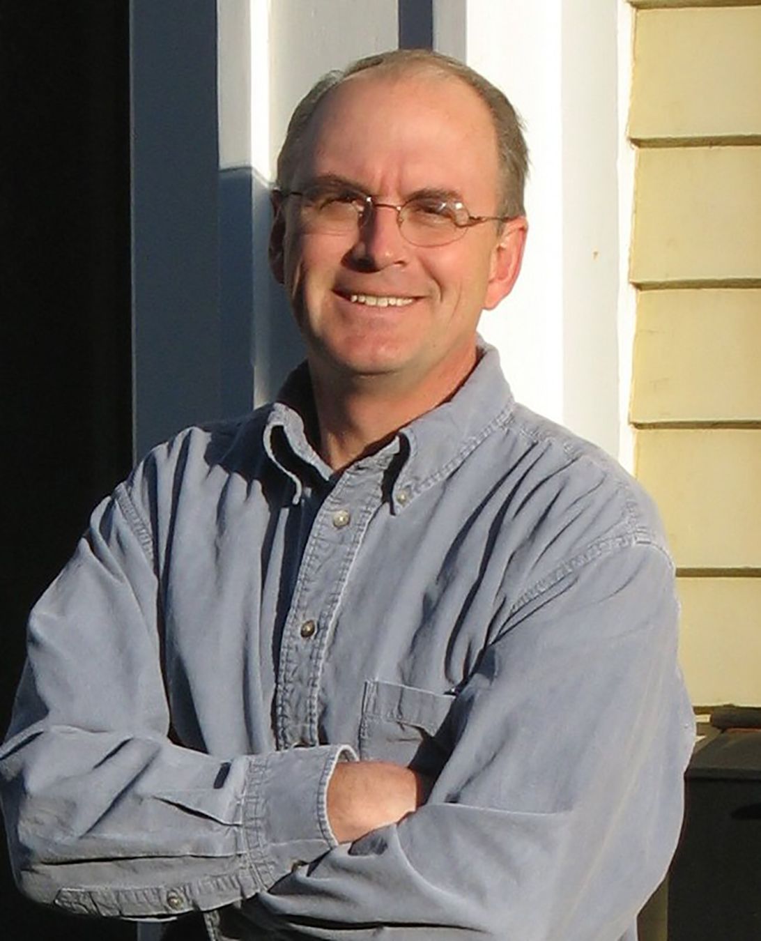 color photograph of a man wearing glasses and a gray shirt lleaning against a door frame with folded arms