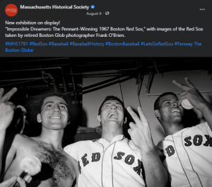 A screenshot of a Facebook post. There is text at the top and an image on the bottom. The image is a black and white photograph of three baseball players from a view around their waists looking up at them. The three men are smiling and looking forward and not at the camera. Two are wearing Red Sox jersey’s, one is bare-chested. They each hold up their index and middle fingers, showing the back of their hands to the camera, the one on the very right is holding a baseball in the same hand. There is a microphone held up below them, close to the camera, and the background has a ceiling and pipes. 