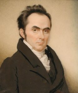 Color photograph of a watercolor painting of a middle-aged man with receding black hair cut short, sideburns and light brown eyes. He wears a white cravat and high collar with a brown jacket. He sits in a three-quarter profile view to the right while looking at the viewer. The background is plain cream that darkens to create a shadow effect behind the sitter.