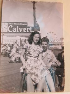 Photograph of Sammy in his uniform in a wheelchair on the boardwalk with a ferris wheel in the background. Bernice is in a dress and sitting on his lap.