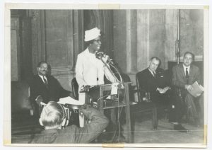 Black and white photograph of a woman in a white dress and hat speaking at a podium. Two men sit behind her to her left and one to her right. In the left foreground, the back of a photographer taking pictures is visible.