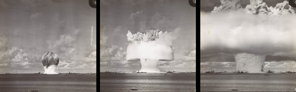 Image of three black and white photographs. The photo on the left shows detonation with a very small cloud where the bottom of the cloud is white and the top is dark over a large body of water; the middle photograph shows a white mushroom cloud over a large body of water; the photograph on the right shows a wide white mushroom cloud that spreads beyond the limits of the photograph over a large body of water