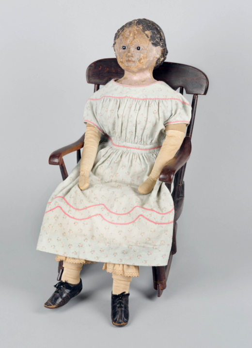 Photograph of a doll sitting in a rocking chair. The doll is wearing a dress and shoes. Her face is cracked and her hair is painted on.