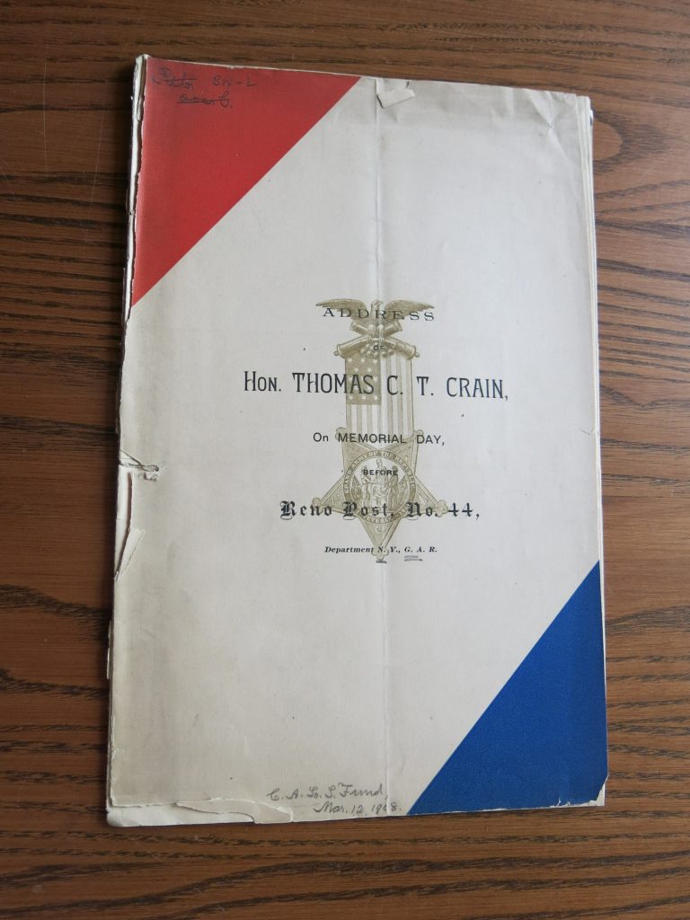 Cover of Thomas Crain’s address. Cover has red, white, and blue stripes diagonally across the cover, starting with red in the left corner. The text is in the center of the white stripe and is black. There is an image of an eagle holding an unfurled American flag that ends in the seal of the Grand Army of the Republic enclosed in a star behind the text. Text reads: Address of/Hon. Thomas C. T. Crain,/On Memorial Day,/Before Reno Post , No. 44,/Department N.Y. G.A.R.