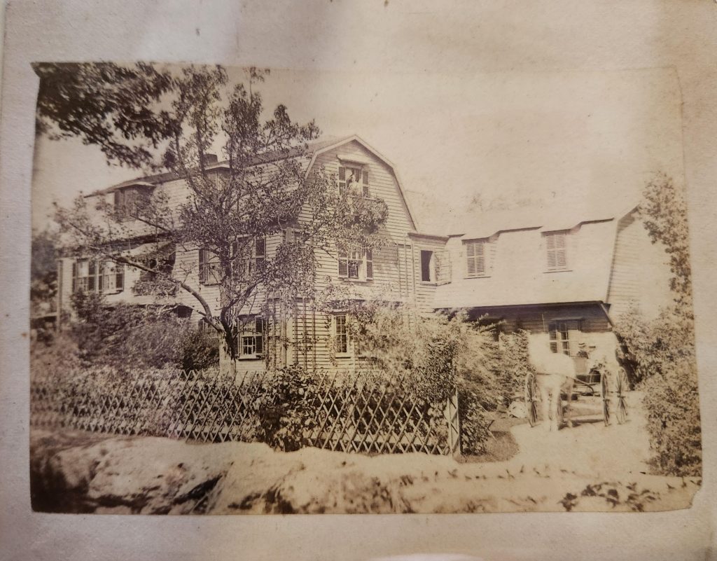 Black and white photograph of a gambrel-roofed three story home with a cross-hatched fence and several trees and shrubs in the yard. A horse and buggy with two figures in the buggy are moving from the driveway to the street. 