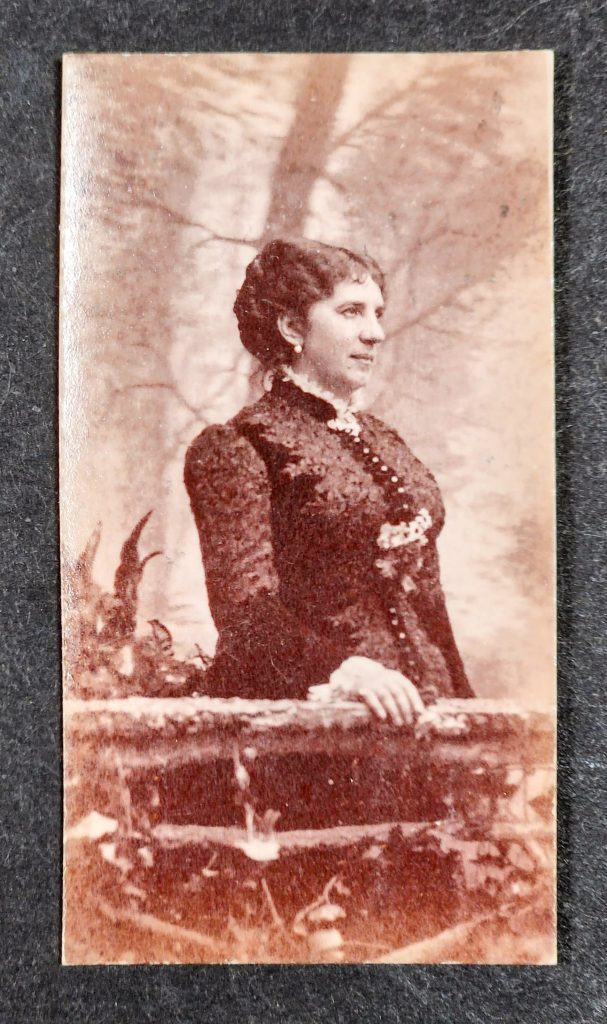 Color photograph of a black and white photograph with a red tint to it. The subject is a middle-aged white woman with dark coiffed hair. She wears a dark dress with buttons running down the front and white lace at collar and cuff. She is looking off to the right. 