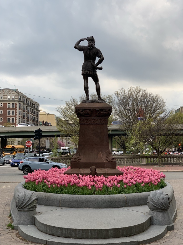 Photograph of bronze statue depicting a man on a stone plinth, taken from the backside view, surrounded by pink tulips. Behind the statue is an overpass with cars