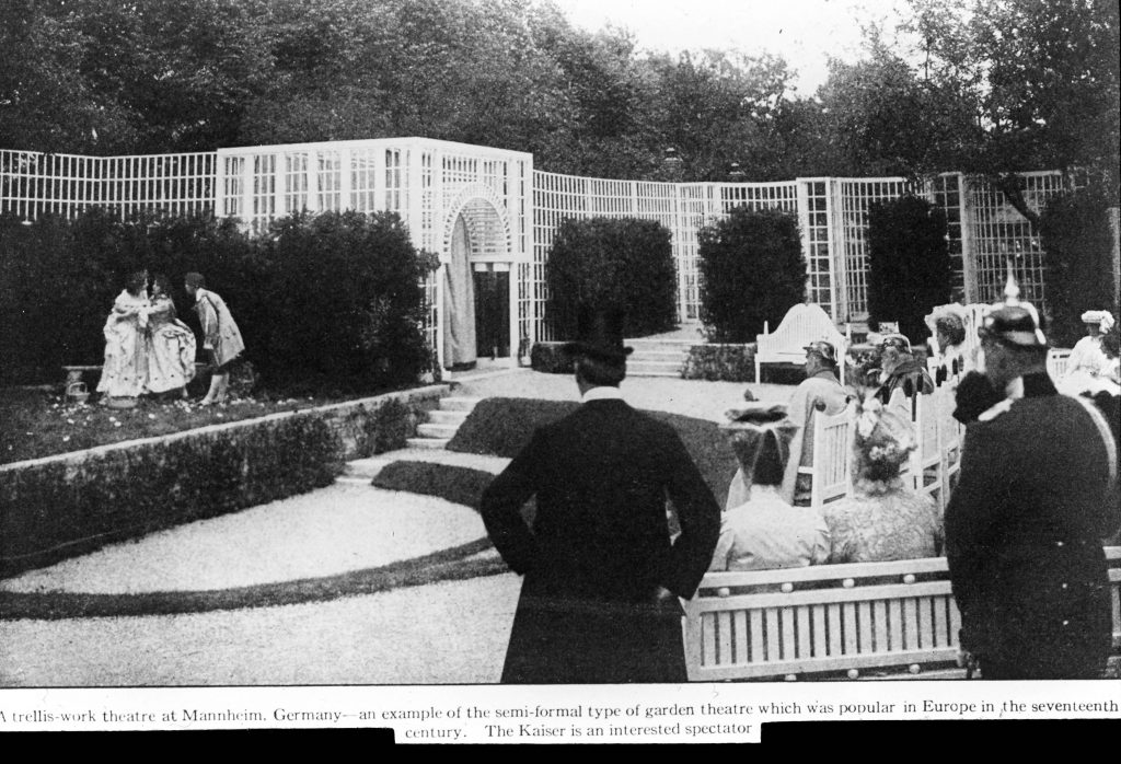 Color photograph of a black and white photograph of an outdoor scene in a garden. There is a raised grassy area to the right with three people in costumes acting out a scene. Behind them are bushes and a long trellis-like fence. To the right are men and women in clothes from the turn of the 20th century (hard to pinpoint a date), some in German military uniform, seated and standing watching the performance. At the bottom is a note that reads "A trellis-work theatre at Mannheim, Germany -- an example of the semi-formal type of garden theatre which was popular in Europe in the seventeenth century. The Kaiser is an interested spectator."