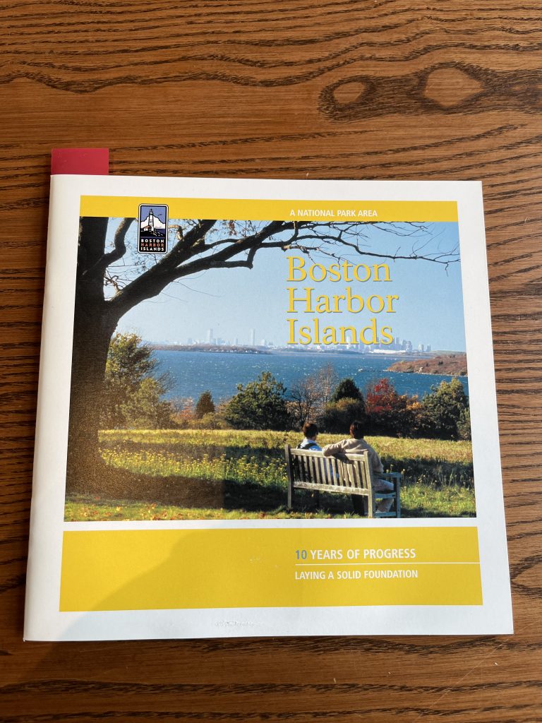 booklet with yellow borders and a picture of two people on a bench looking out onto the water with “Boston Harbor Island” written in yellow on the top right of the picture. In the bottom right corner of the booklet, it reads “10 years of progress/Laying a solid foundation” in white.]