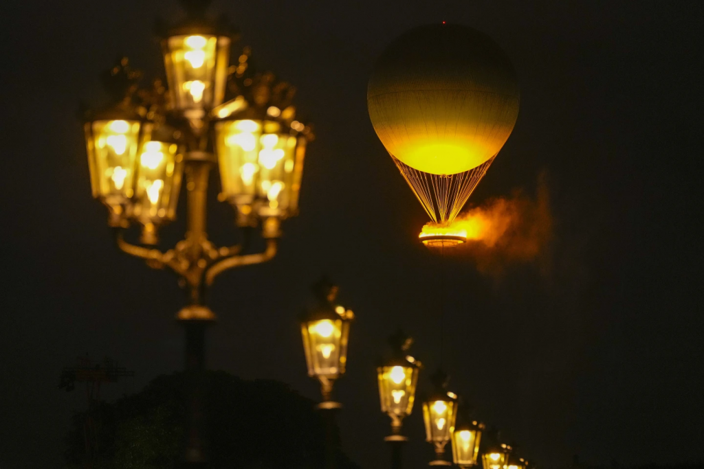 Color photograph aimed upwards from the ground on a dark night. What is visible is a large inflated balloon tethered by many strings to a basket holding light and giving off steam which lights the balloon from below with a lovely glow. A row of old-style streetlamps are in a line from the top left to the bottom right lit up to match the balloon. 