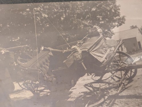 Color photograph of a black and white photograph of a black horse-drawn carriage with mangled wheels on the side closer to the camera. 