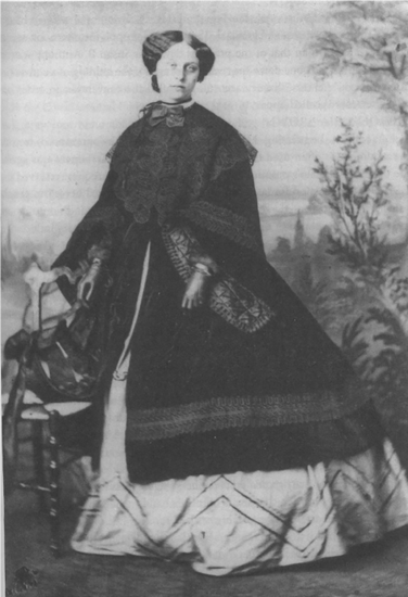 Black and white photograph of a young white woman in a large-skirted light-colored dress with a black jacket draping far down the skirt and with wide sleeves. Over that is a black lace shawl that covers her shoulders. Her hair is pulled back but there is a waviness to her dark hair that shows. She stands by a chair and the background is a bush and tree that looks painted like a background. 