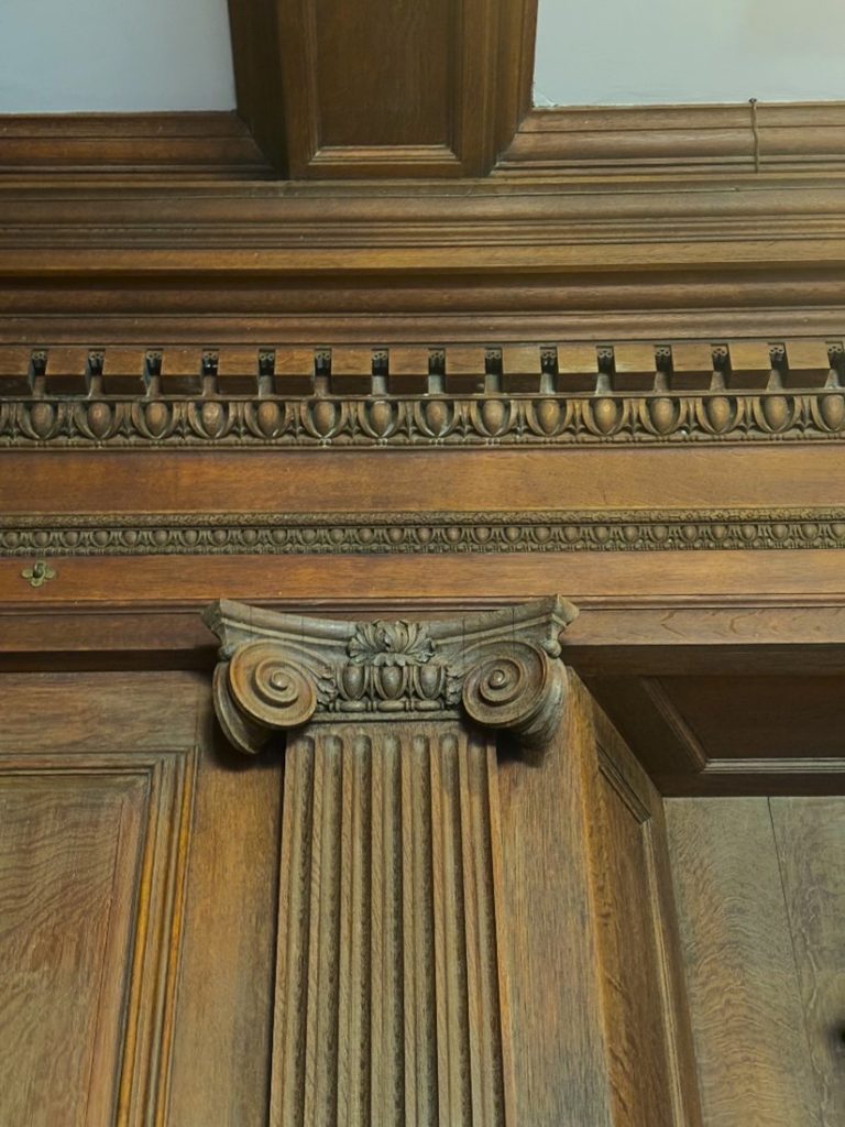 Close-up of a wooden wall section. A fluted pilaster and the decorative bands of the entablature are centered in the frame.