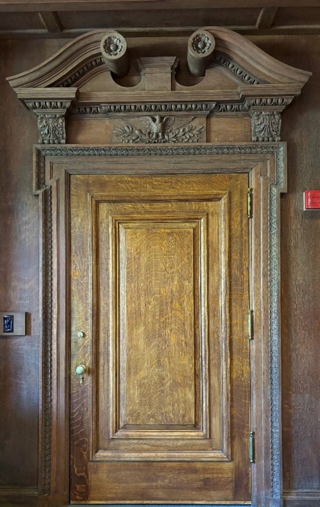 A wooden door in Ellis Hall. It is richly decorated on top, with a frieze, carving of an eagle, and swan's necks pediments curling near the ceiling.