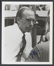 small black and white image of a white man with thinning hair wearing a white shirt with a dark tie. He is wearing large glasses on his face and is frowning.