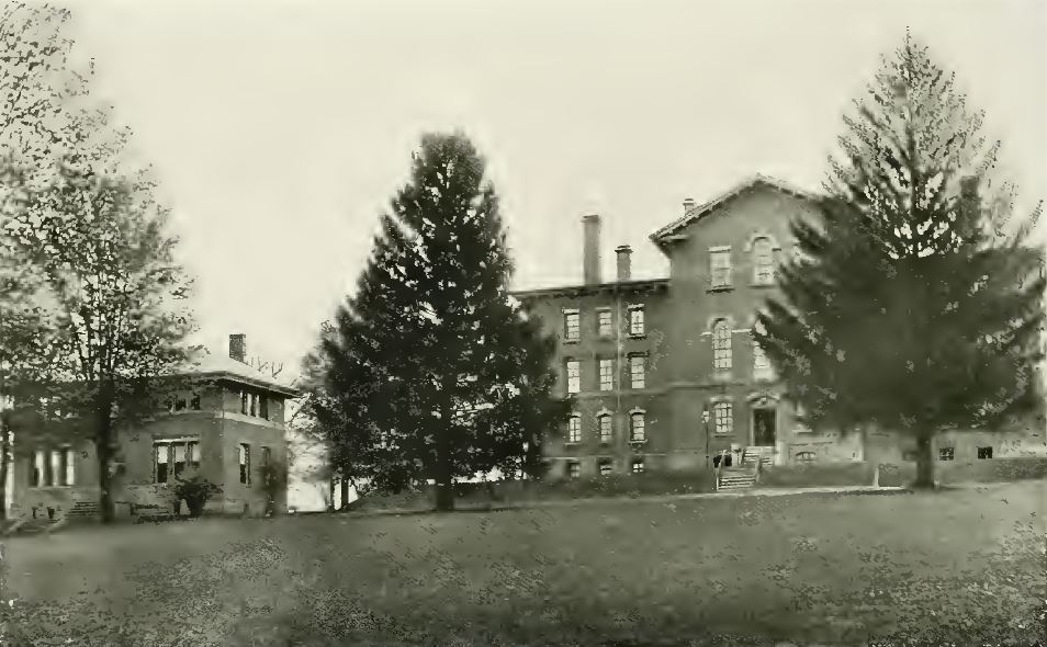 Black and white photograph of two buildings, a three-story building on the right and a smaller two-story one on the left. In front of the buildings is a large lawn and three trees.