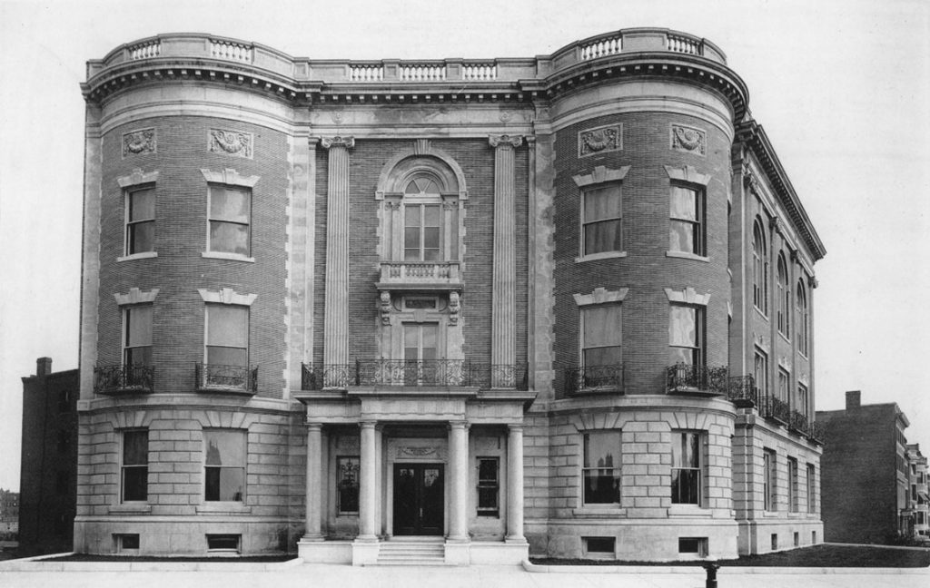 A black and white photo of the large, sturdy Massachusetts Historical Society building. A few small buildings are visible in the background.