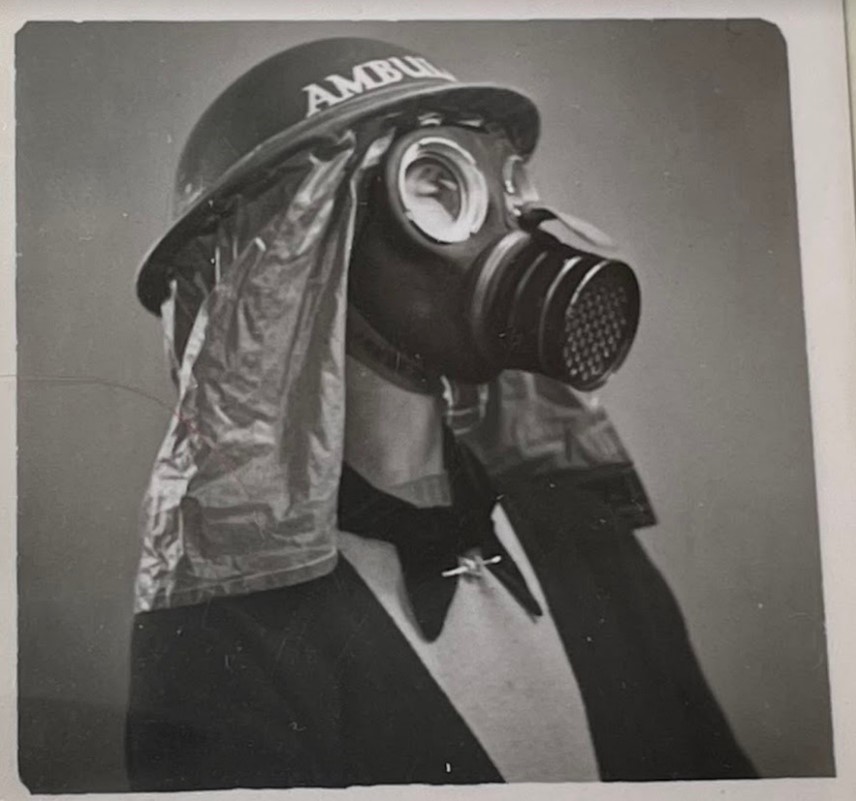 Black and white photograph of a white woman in a 1940s style gas mask. She is wearing a black jacket, light sweater, and a black tie. 