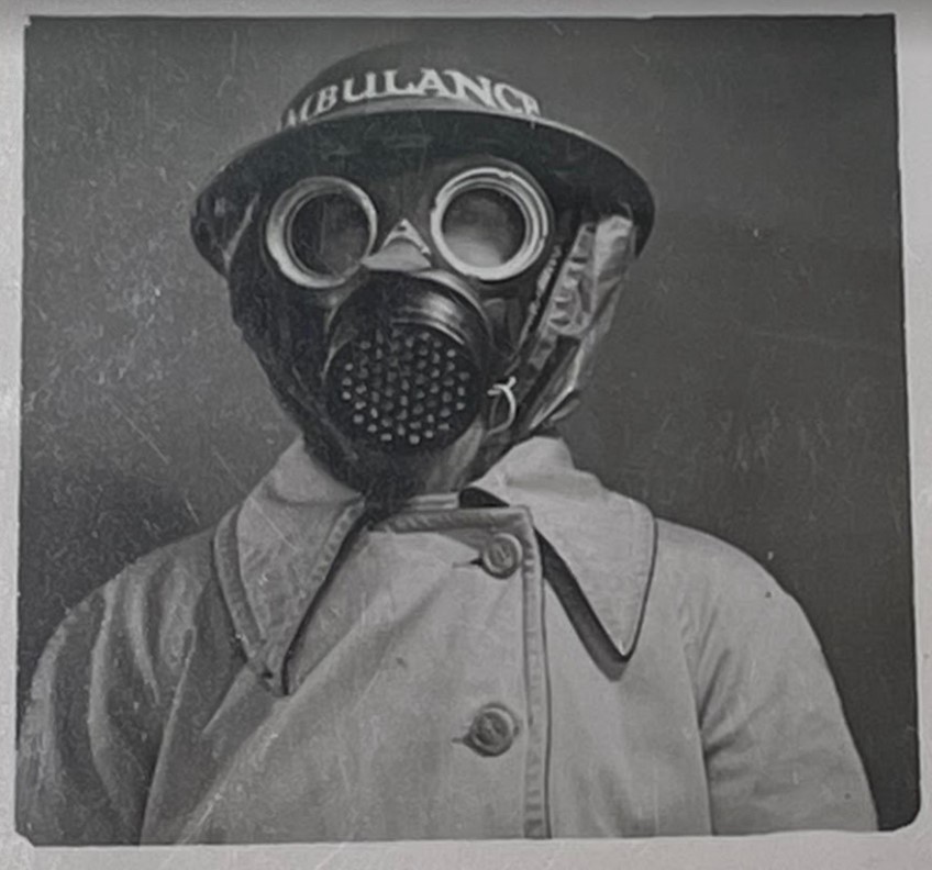 A black and white photograph of a woman wearing a 1940s style gas mask. She is wearing a light-colored buttoned-up trench coat. 