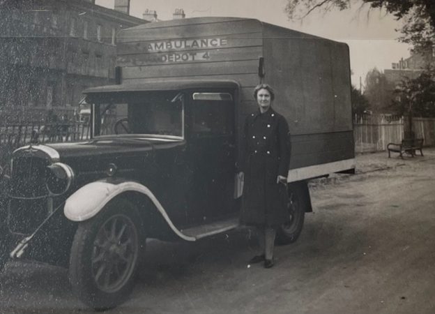 Black and white photograph of a middle-aged white woman standing next to a 1940's truck with "Ambulance, Depot 4," written on the wooden extension over the bed of the truck. She is wearing a long dark coat. 