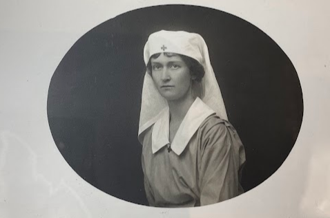 Color photograph of a black and white photograph in an oval shape. The image is of a young white woman with dark hair under a nurse's white habit with a medical cross at her temple. She wears a smock style dress with a wide white collar. The background is very dark. 