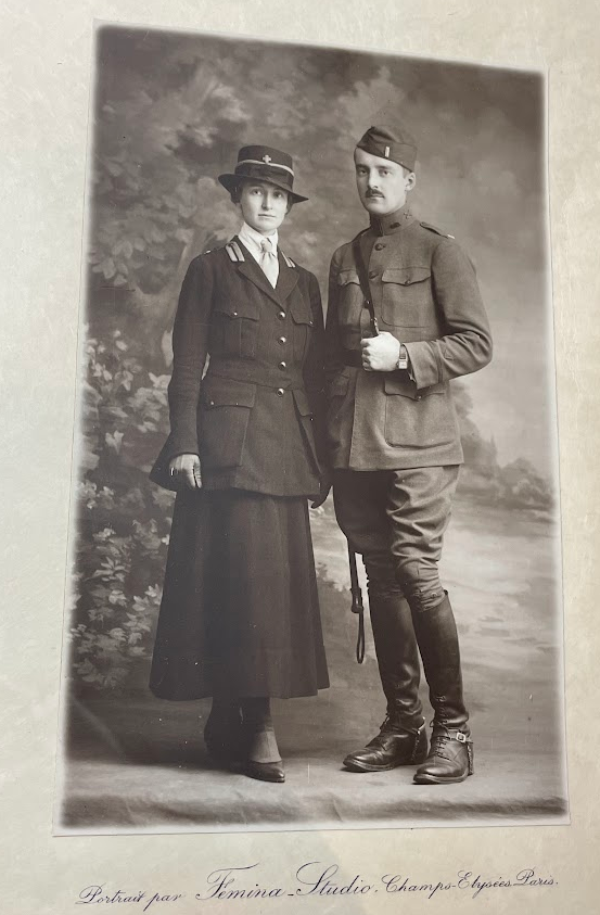 Color photograph of a black and white photograph of a man and a woman standing together in front of a painted backdrop of trees and a field. She stands to the left in a military red-cross uniform, a long skirt, flared jacket, white shirt and light tie and a hat with a brim and the Red Cross cross on it. To the right the man is in a WWI military uniform with high leather books, flared pants at the hips - tight at the knee, a long military jacket with leather belt and shoulder strap and hat. 