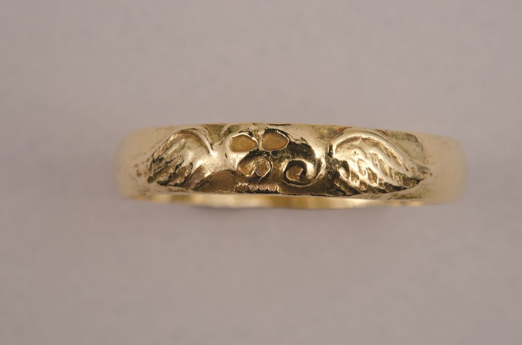 A color photograph of a gold ring with a skull with wings on either side pressed into it. 