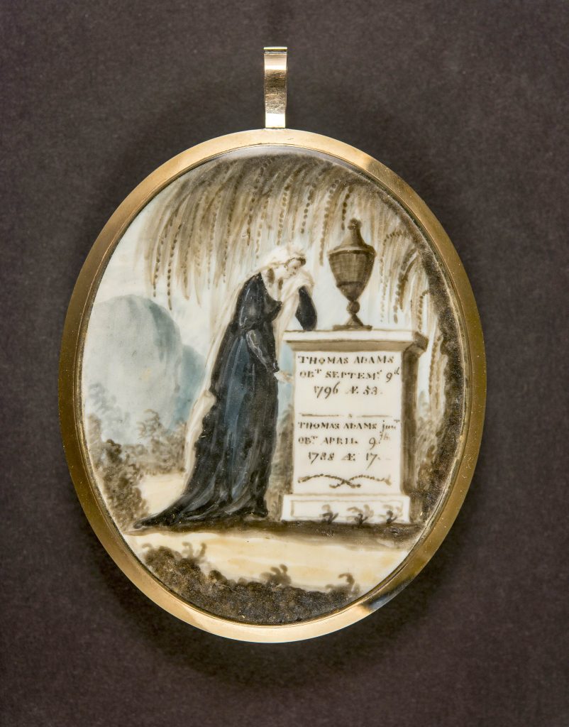 A color photograph of a painted image in an oval with gold metal around it making it into jewelry. The painted image is of a woman crying over a gravestone with an urn on top and a weeping willow tree above her. The gravestone has writing on it for Thomas Adams.  