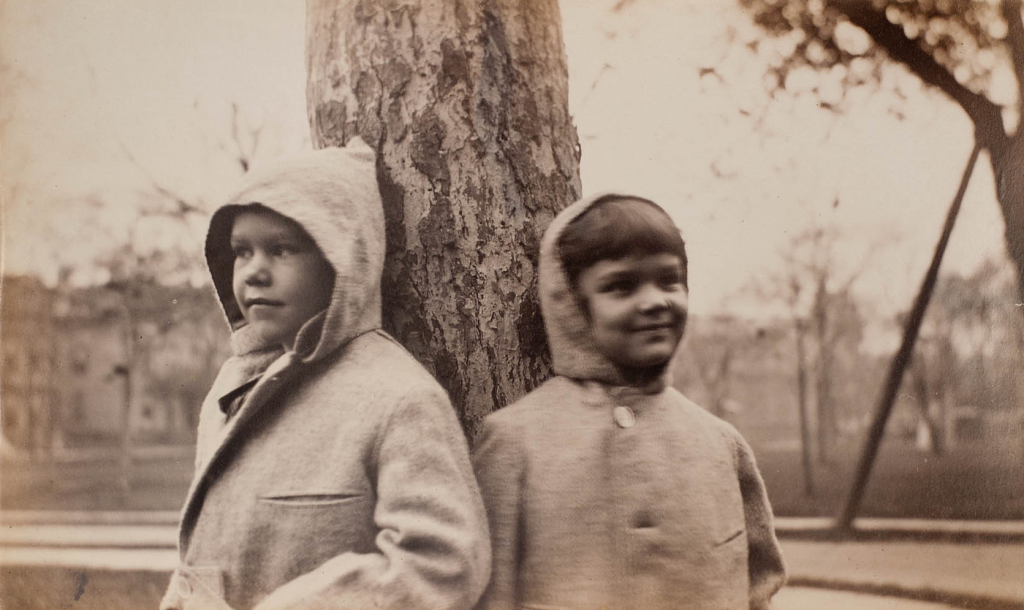 Sepia tone photograph of two children in hooded coats with the hoods up standing with their backs to a tree in a field. Trees and a house are out of focus in the background and each child looks off into the distance on their respective side, left and right. 