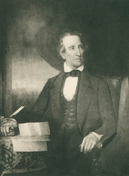 A white man in a suit sits at a desk strewn with papers. His hand is on a book.