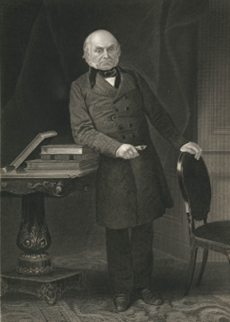 A white man in a long coat stands next to a desk.