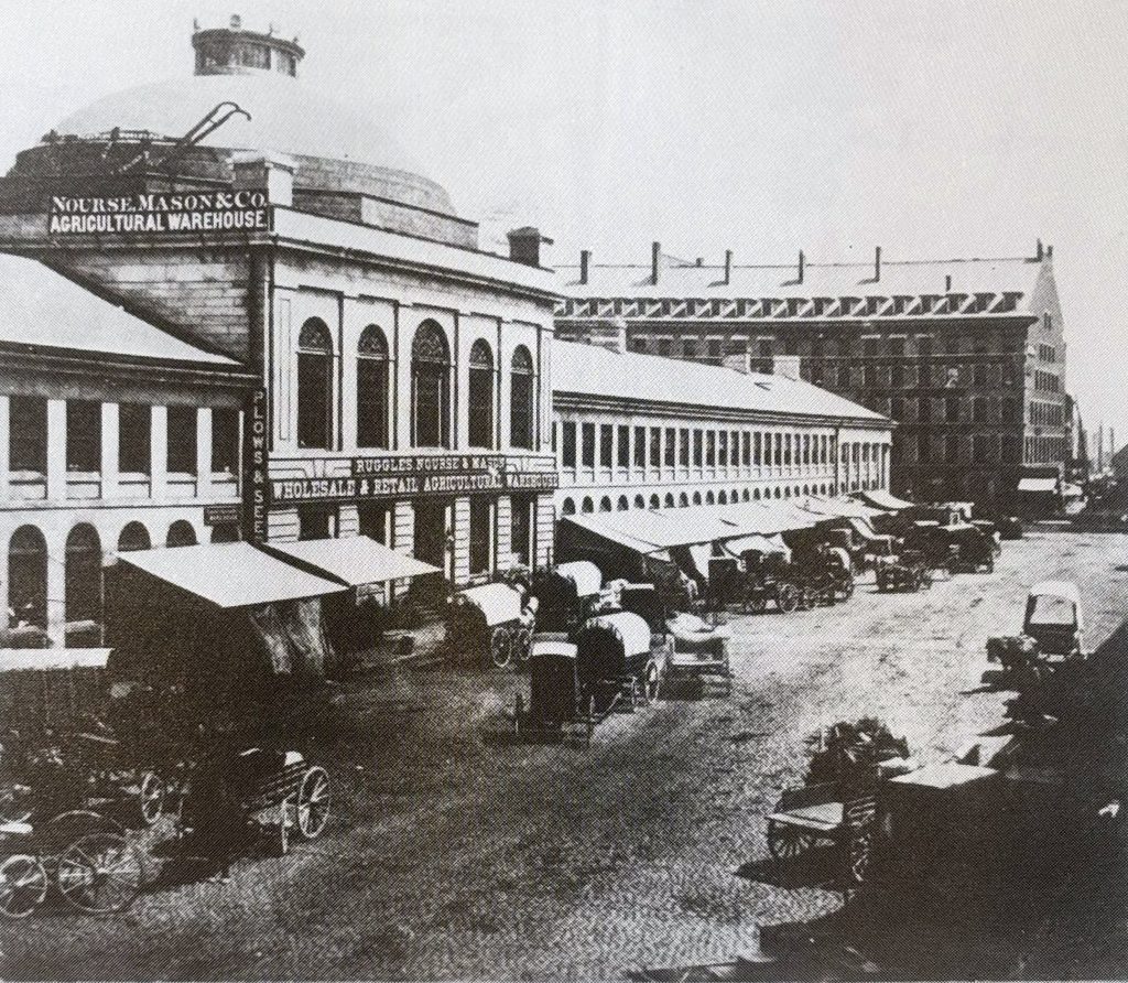 Next to the large stone side of Quincy Market, a street with market tents and covered wagons.