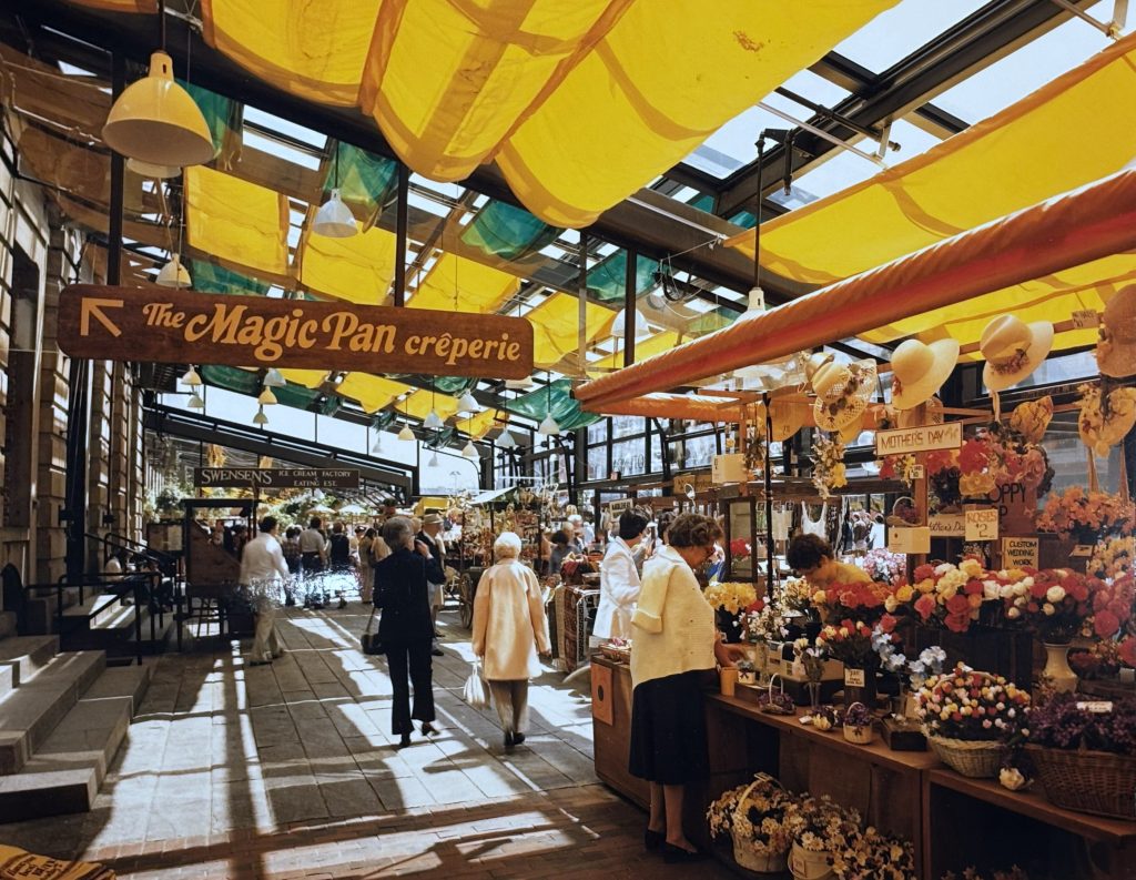 Shoppers browse market stalls selling flowers and other goods under colorful sun shades. A sign for "The Magic Pan Creperie" points away.