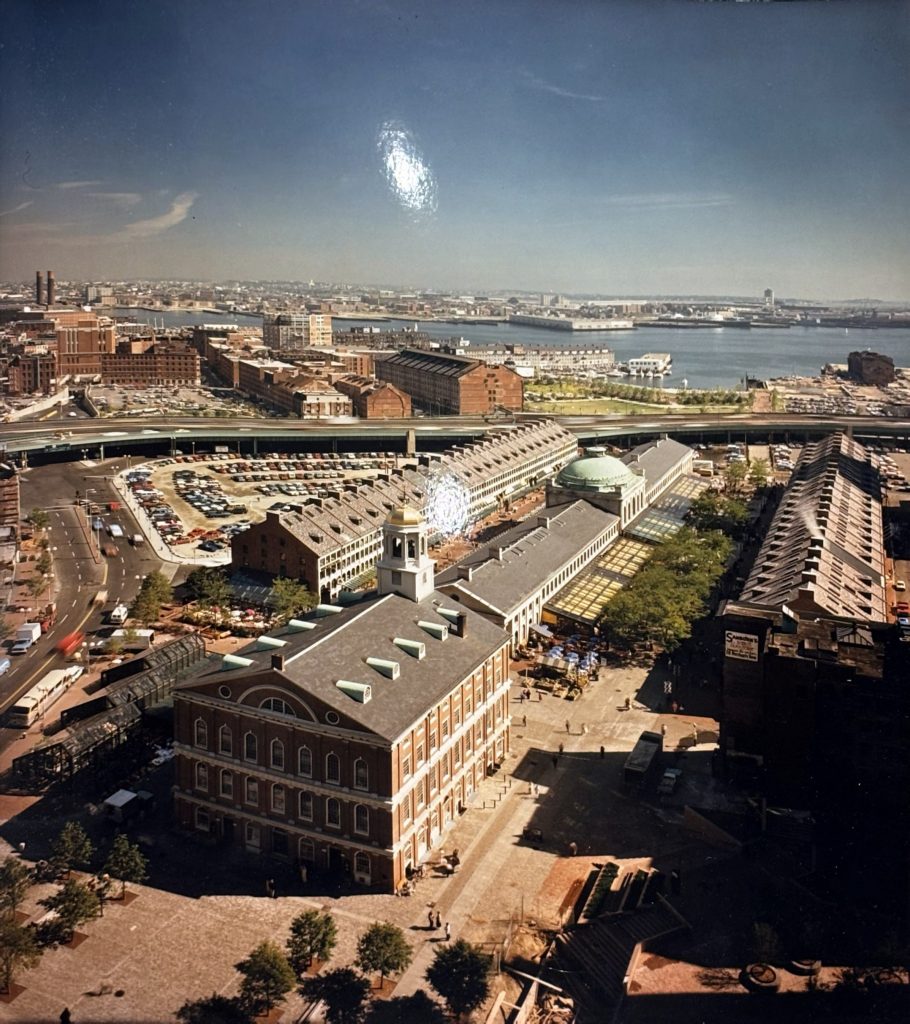 An aerial view of Faneuil Hall and Quincy Market. Pedestrians walk around the buildings, while roads and highways stretch to the sides and rear of the marketplace. Boston Harbor is visible in the background.