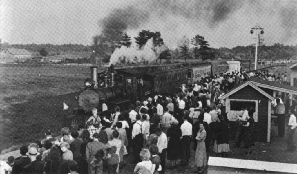 Black and white photograph of a crowd of people standing outside next to a train pulling into a station. The train engine pulls several cars, and smoke billows out of its chimney. Behind the train and at the left of the picture are fields, trees, and farm buildings. At the right is a small station building and a railroad signal on a pole.