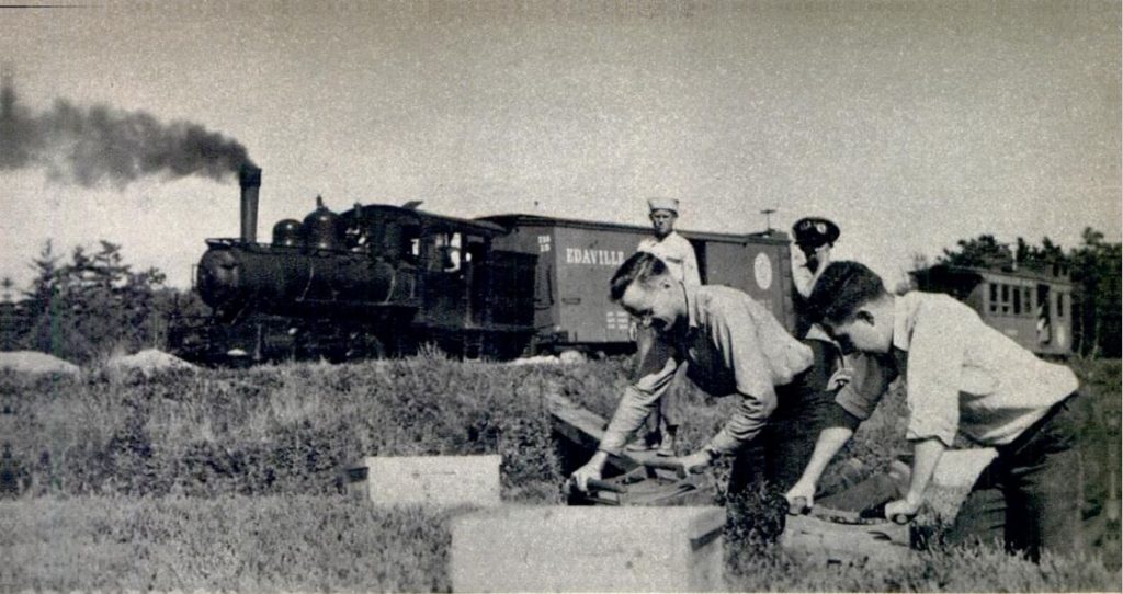 Black and white photograph of workers in a field with a train passing behind them. In the foreground, two kneeling workers are bending over and holding onto wooden crates. Two other workers stand behind them and watch. The train in the background has smoke billowing out of its chimney and the word “Edaville” written on its side.