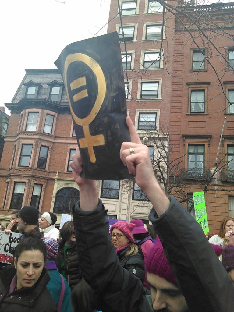 Photograph primarily depicts a sign held up by a protester in a crowd of people. The sign features a woman’s symbol with an equals sign.  Brick and sandstone buildings are in the background.  