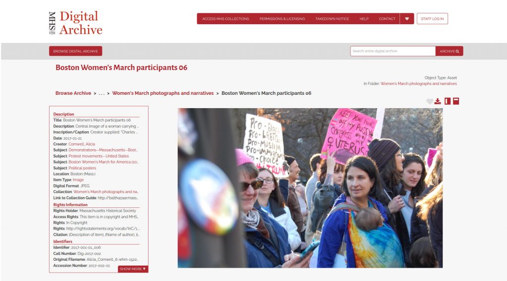 Screenshot from the MHS Digital Archive showing Boston Women’s March participants image 06.  A column of metadata is on the left.  The image depicts a large crowd of people standing close to each other, two people are holding signs.  A woman on the left appears to be looking at her cell phone. A woman in the middle of the photograph is looking to the left at various people and she is carrying an infant in a colorful tie-dye sling.  