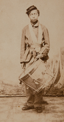 <p>A black and white photograph of a young, Black boy wearing a cap and a large army coat cinched at the waist with a belt standing against a white background. He holds one drumstick in each hand, one of which is resting against a large snare drum which hangs from his neck.</p>