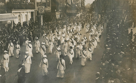 A black and white photo of a number of women marching in the street, wearing matching white outfits and dark sashes while crowds look on